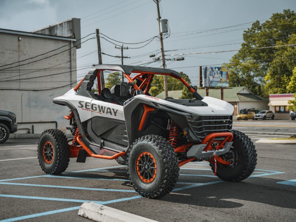 Off-road Segway vehicle in a parking lot with an orange frame and white body, labeled 'SEGWAY' on the side, large knobby tires visible.