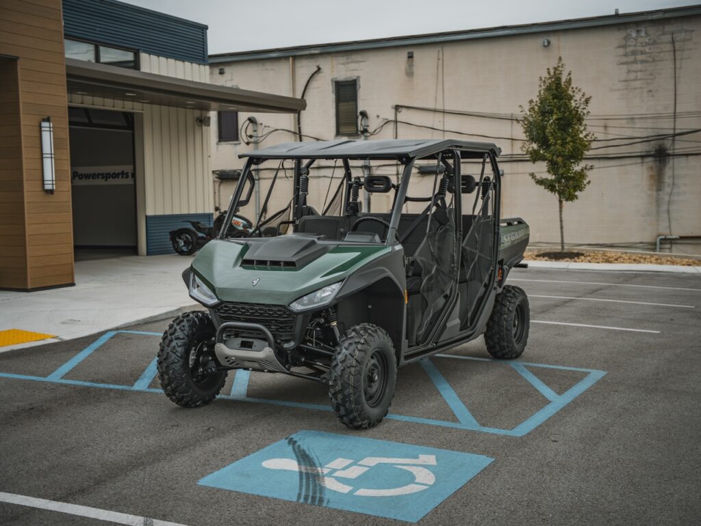 Green off-road utility vehicle (UTV) parked in a handicapped parking space outside a building. Includes roll cage and four seats ahead of the dealership area.
