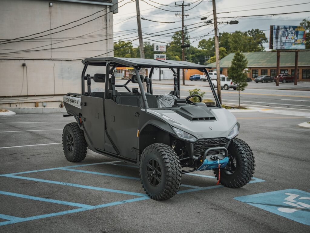 Gray open-top off-road utility vehicle with roll cage and large knobby tires parked in a handicap parking space in an urban lot.
