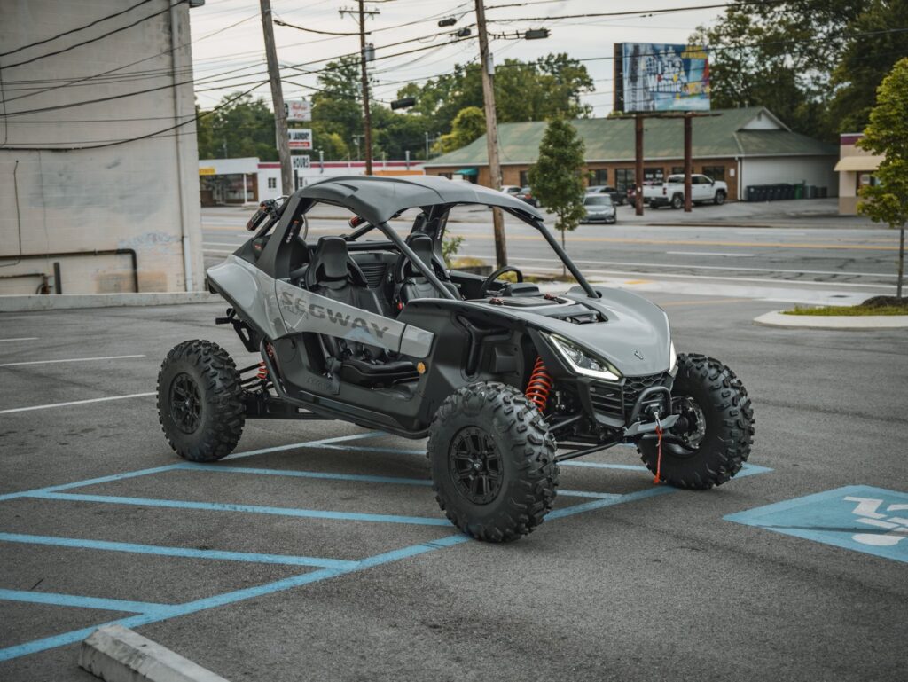 Gray off-road side-by-side UTV parked in a blue handicapped parking space, urban backdrop behind it, roll cage visible.