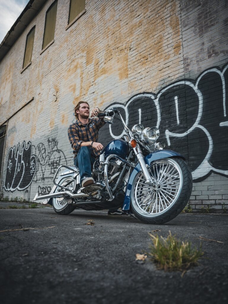 Man in a plaid shirt sits on a blue cruiser motorcycle with chrome detailing beside a graffiti-covered brick wall.