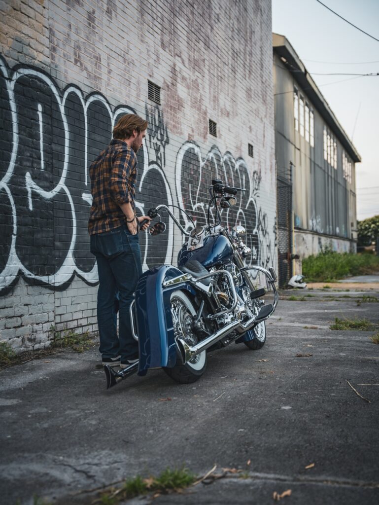 Man in a plaid shirt stands beside a blue motorcycle parked against a graffiti-covered brick wall in an urban alleyway
