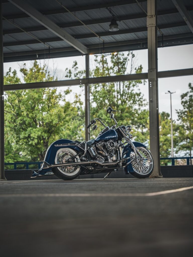 Blue custom motorcycle with chrome accents parked under a metal canopy, trees visible in the background outside the structure.